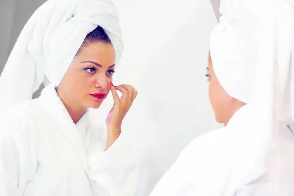 Young woman with a towel wrapped around her head examining her skin in the bathroom mirror as she looks for blemishes with a thoughtful expression