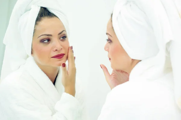 Young woman with a towel wrapped around her head examining her skin in the bathroom mirror as she looks for blemishes with a thoughtful expression