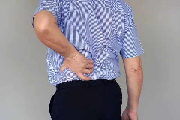 Close-up elderly man with pain in kidneys isolated on a gray background. Old man with back ache clasping her hand to her lower back. Man suffering from ribbing pain, waist pain.