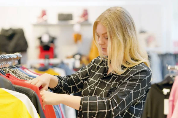 Young blonde girl, choose new things in the store. Beautiful young female shopper chooses liked things in the boutique. A young girl chooses things on a hanger in a store.
