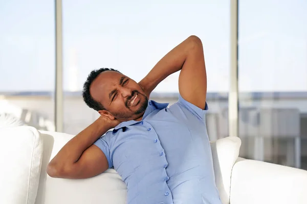 Tired neck. Man suffering from neck pain at home on couch. Males sense of fatigue, exhausted, stressed. African man massages her painful neck with her hands. The concept of body and health.