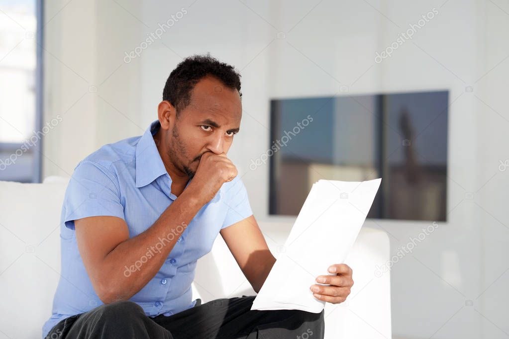 Man reads negative news in a letter at home on the couch. The shaken business manager of the african male received a notice of dismissal from the company, surprised. An agitated man without joy.
