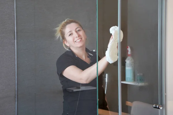 La femme de chambre dans la salle de bain essuie le verre de la salle de douche dans la chambre d'hôtel. Nettoyage. Femme nettoie le verre dans la salle de bain — Image Femme Chambre Dans Salle Bain Essuie Verre Salle Douche Dans Photo De Stock