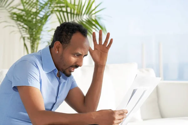 Man reads negative news in a letter at home on the couch. The shaken business manager of the african male received a notice of dismissal from the company, surprised. An agitated man without joy.