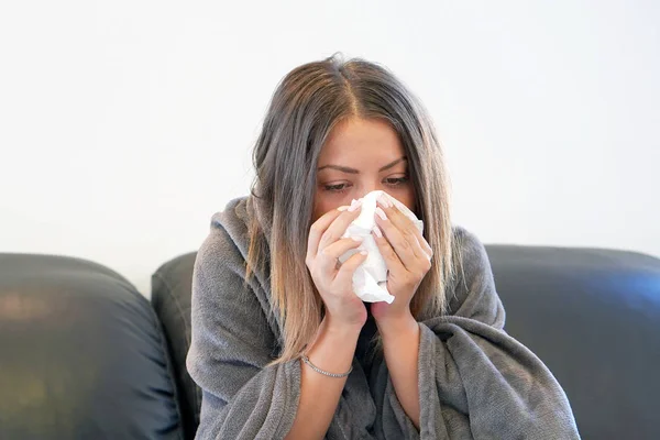 Attractive woman feeling cold, freezing, with napkin in a hand, wrapped in blanket, sitting on the sofa. Unhappy upset tired woman is sitting on a sofa at home suffering from a cold and using napkins.