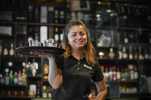 Garson, bardaki otel restoranının tepsisinde viski bardağı taşıyor. Hizmet kavramı. Arka planda alkol şişeleri olan raflar. Gece vakti..