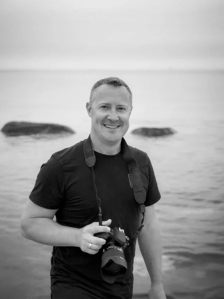 a male smiling photographer on tour on the beach to search for motives with a camera around his neck and in his hand in a black polo shirt with a view into the camera