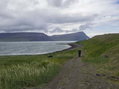 İzlanda, Batı fjords, Hornstrandir, Latrar, 26 Haziran 2018: patika iz adalvik Cove doğa rezerv Hornstrandir, yazlık evler, yeşil çim çayır, taş beach ile okyanus, yamalı kar cliff yürürken ağır sırt çantası ile yalnız adam uzun yürüyüşe çıkan kimse