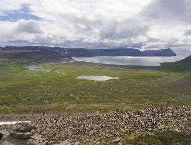 Adalvik ve latrar göl ve nehir akarsu, yeşil çim çayır, plaj, okyanus, tepeler ve dramatik uçurumlar, karanlık bulutlu gökyüzü arka plan ile Batı fjords doğa rezerv İzlanda'daki Hornstrandir görüntüleyin