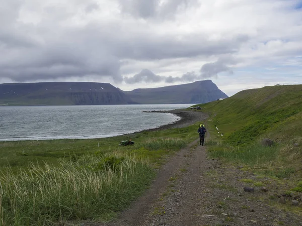 アイスランド 西フィヨルド ホットンストランディル Latrar 2018 孤独な男の重いバックパックをパッチ雪崖 海自然保護区ホットンストランディル 夏のコテッジ 緑の草の牧草地 石のビーチで — ストック写真