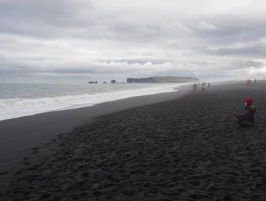 South Iceland, Vik ben Myrdal, 4 Temmuz 2018: genç güzel kadın meditasyon ya da Reynisfjara siyah kum plaj, Dyrholaey rock ve grup backgrund, sis ve huysuz gökyüzü, tek renkli göz turist insan üzerinde rahatlatıcı oturan kırmızı şapkalı