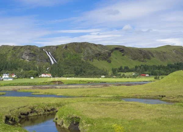 rural landscape with colorful farm houses, blue river stream on green grass meadow with spruce tree forest, hills and beatuful waterfall Systrafoss in Kirkjubeajarklaustur