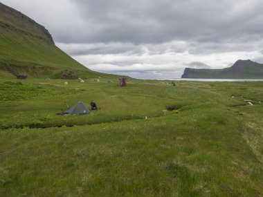 Iceland, west fjords Hornstrandir, June 29, 2018: Hornvik campsite with view on beautiful Hornbjarg cliffs in remote nature reserve with green meadow, water stream and hills, blue sea and cloudy sky background. clipart