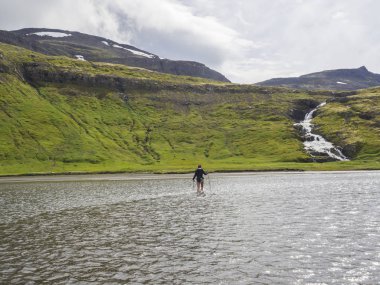 Uzun yürüyüşe çıkan kimse kadın ceket, mor şapka ve çıplak bacaklar arka planda Hornbjarg cliff, şelale ve yeşil Hills Trek'te İzlanda Hornstrandir Nehri geçerken trekking Polonyalılar ile. Yürüyüş ve eğlence Tema