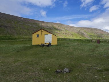 Adalvik koyu sarı acil barınma kabin içinde Batı fjords İzlanda, Hornstrandir yeşil çim çayır, tepeler ve mavi bulutlar arka plan ile kamp yeri latrar görüntüleyin