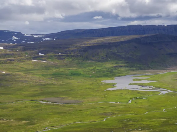 Vista sobre hermosos acantilados en la cala adalvik en la reserva natural de fiordos del oeste Hornstrandir en Islandia, con delta del arroyo del río, casa roja, prado verde cubierto de nieve y fondo de nubes oscuras —  Fotos de Stock