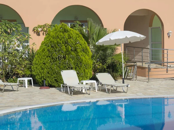 Empty hotel swimming pool during sunny day, summer vacation. View of blue water in pool, white sunbeds, umbrella and green palms, Greece, Corfu island — Stock Photo, Image