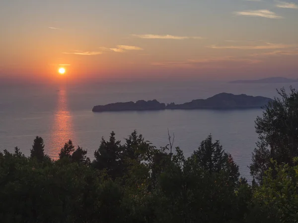 Όμορφη πολύχρωμα πορτοκαλί ηλιοβασίλεμα ήλιο να πέφτει στον ωκεανό με τα βράχια στη Μεσόγειο θάλασσα στο Πόρτο Timony, πράσινο θάμνο σε πρώτο πλάνο Κέρκυρα, το νησί — Φωτογραφία Αρχείου