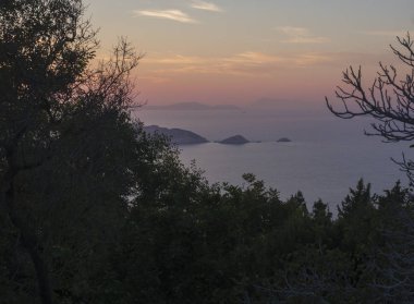 Güzel renkli turuncu günbatımı gökyüzü, ön plan Corfu Island, Yunanistan ile Akdeniz kayalarda Porto Timony, yeşil bush toprak çıplak ağaca üzerinde okyanus şubeleri.