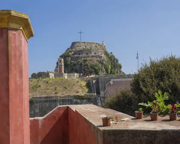 Pared roja de la iglesia con vista al famoso monumento turístico antigua fortaleza veneciana con torre del reloj, ciudad de Kerkyra, Corfú, Grecia, día de verano, fondo azul del cielo — Foto de Stock