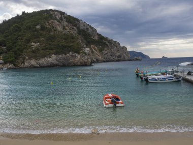 Yunanistan, Corfu, Paleokastritsa, 26 Eylül 2018: kırmızı motorbot ve Paleokastritsa defne bech, yeşil tepe ve deniz, iskelede otopark turist tekneleri Yaz bulutlu gökyüzü, Corfu, Yunanistan