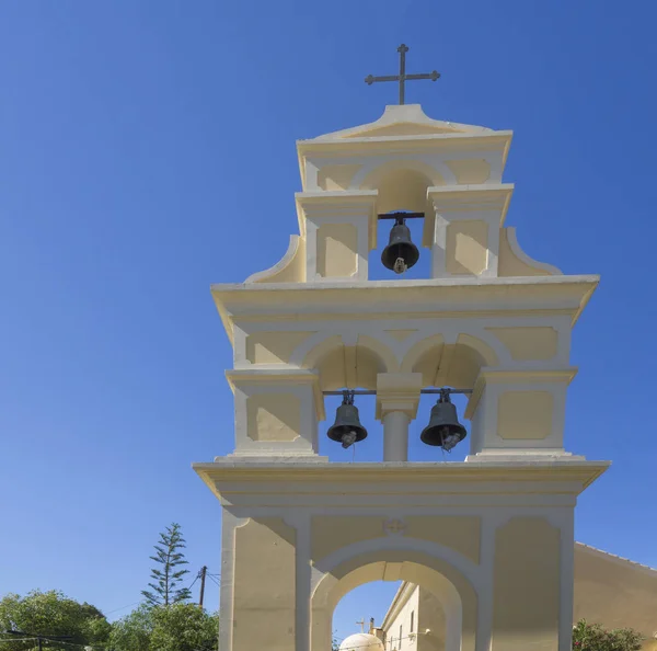 Traditional Greek Freestanding bell tower at church in Sidari, yellow white belfry with three bells and cross at Sidari, Corfu Greece, blue sky background.
