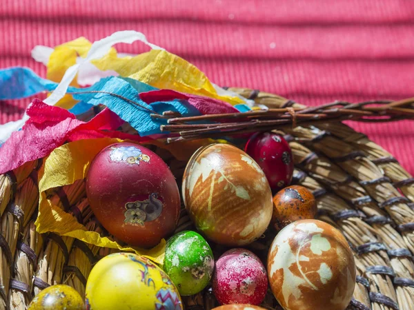 Homemade Colorful Painted easter eggs in straw flat basket with easter stickers and Pomlazka - czech traditional braided whip made from pussywillow twigs on red tablecloth background, selective focus