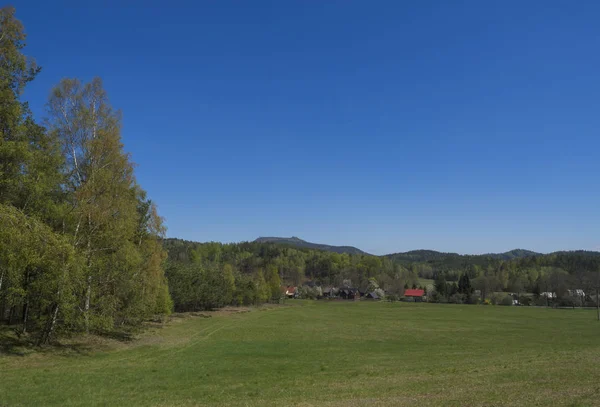 Paesaggio primaverile con vista sul villaggio di Marenice nelle montagne lusiziane con cottage in legno tradizionale e prato di erba verde lussureggiante, betulla latifoglie e abete rosso foresta e colline, sfondo cielo blu — Foto Stock