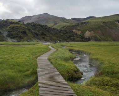 Landmannalaugar kamp alanında bir termal banyolarda dinlenen turist grubu ile doğal kaplıca ahşap yol, İzlanda. Arka planda çayır, lav tarlaları ve dağlar