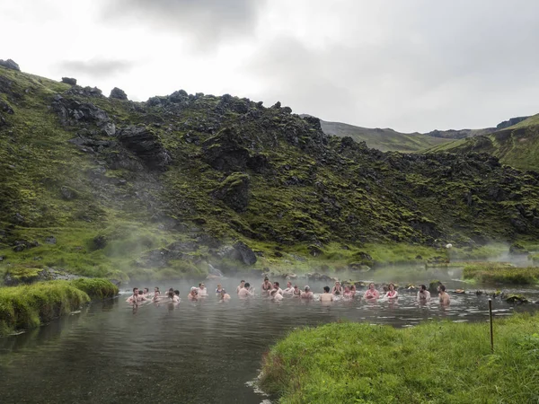 Islândia, Landmannalaugar, 30 de julho de 2019: Grupo de turistas relaxando em uma fonte termal natural em banhos termais no acampamento Landmannalaugar, Islândia. Prado de grama, campos de lava e montanhas em — Fotografia de Stock