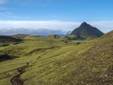 Volcanic landscape with footpath of Laugavegur trek and green Storasula mountain with lush moss and low clouds. Fjallabak Nature Reserve, Iceland. Blue sky background, copy space.