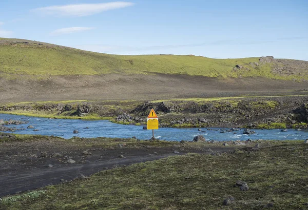 Iceland, South Highlands, August 1, 2019: blue glacier river cross on mountain road F210 with warning notice text crossing requires caution sign, green hills and blue sky background — Stock Photo, Image