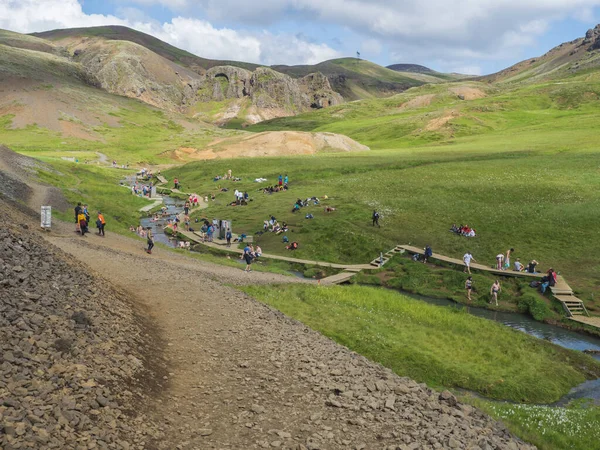 Islandia, Hveragerdi, 5 de agosto de 2019: Baño geotermal natural abarrotado en un arroyo del río caliente en el valle de Reykjadalur con grupo de personas que se bañan y relajan y turista, sendero de madera y cambio — Foto de Stock