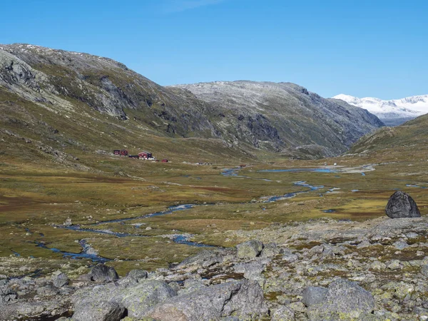 Őszi kilátás a piros Krossbu turisztikai állomás hegyi kunyhó épületek gyalogos gleccser Smorstabbreen kék patak patak, hófödte hegyek és narancs moha és heath a Jotunheimen Nemzeti — Stock Fotó