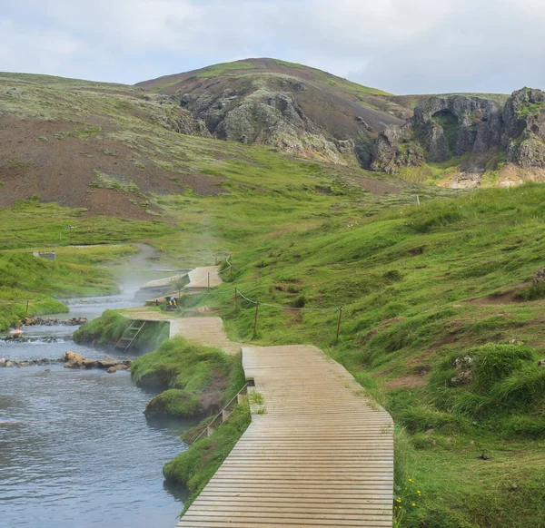 Naturalna kąpiel geotermalna w gorącym strumieniu rzeki w dolinie Reykjadalur z drewnianą ścieżką i przebieralniami. Południowa Islandia w pobliżu miasta Hveragerdi. Letni słoneczny poranek — Zdjęcie stockowe