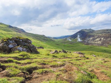 Sıcak pınarlı Reykjadalur vadisi, yemyeşil çayır, jeotermal buharlı kayalar ve tepeler. Güney İzlanda, Hveragerdi şehri yakınlarında. Yaz güneşli sabah, mavi gökyüzü.