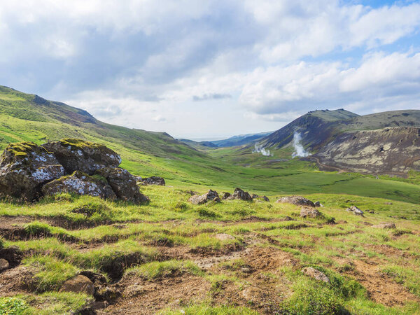 Reykjadalur valley with hot springs river, lush green grass meadow, rocks and hills with geothermal steam. South Iceland near Hveragerdi city. Summer sunny morning, blue sky.