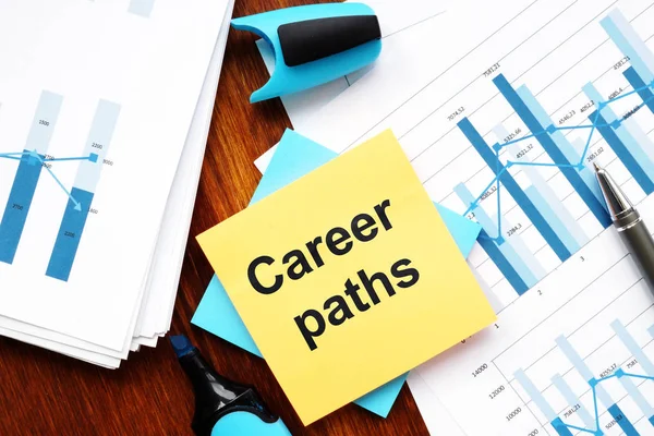 Writing note showing Career paths. The text is written on a small colored paper. Conceptual photo shows papers with graphs and colored papers, markers, wooden background.