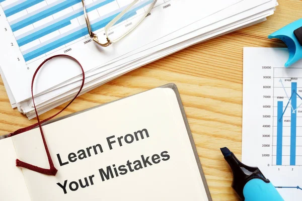 Writing note showing Learn From Your Mistakes. The text is written on a paper of notebook. Papers with graphs and colored papers, markers, wooden background are on the photo too.