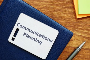 Communications Planning.The text'i gösteren yazı, ünlem işareti silueti olan küçük ahşap bir tahtaüzerine yazılmıştır. Fotoğrafta kitap, kalem, renkli kağıtlar var..