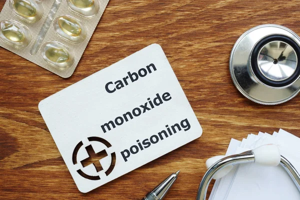 Text sign showing Carbon monoxide poisoning. The text is written on a small wooden board with red cross silhouette. There are blister, pills, stethoscope, papers, wooden table on the photo.