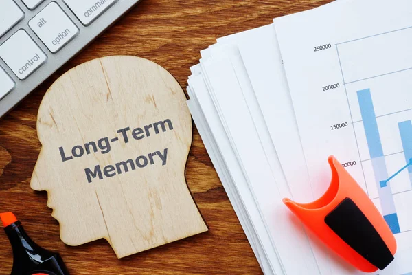 Writing note showing Long-Term Memory. The text is written on a head-shaped plaque. Papers with graphs, keyboard, notebook, a pen are on the photo too.