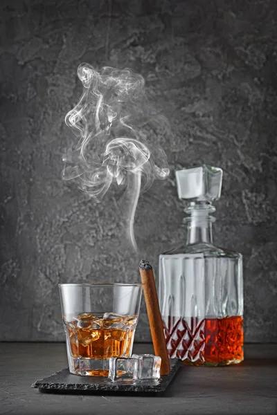 Strong alcoholic drink scotch whisky with ice cube in old fashion glass and decanter with smoking cigar on gray concrete background