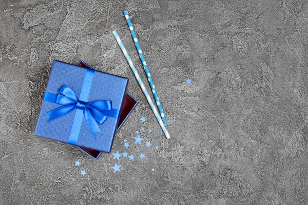 青い光沢のある古典的なギフト ボックスのサテンの弓とコンクリートの灰色の背景にパーティの属性として星の形の紙吹雪が紙カクテル ストロー — ストック写真
