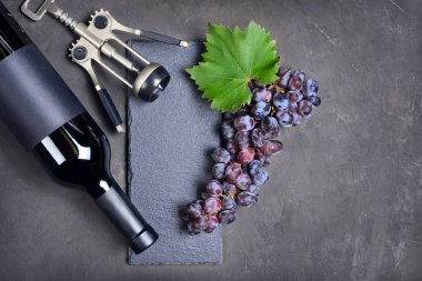 Düz koyu gri beton zemin üzerine taze üzüm ve kopya alanı ile yalancı şarap şişesi, metal tirbuşon ve siyah arduvaz kurulu göklere