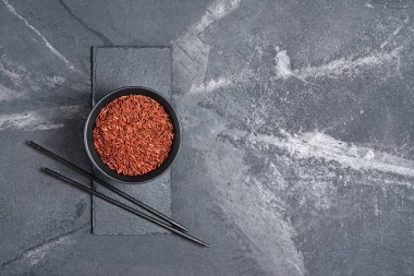 Kırmızı pişmemiş uzun taneli pirinç kayrak kurulu ve siyah mermer zemin ahşap yemek çubukları ile karanlık kase