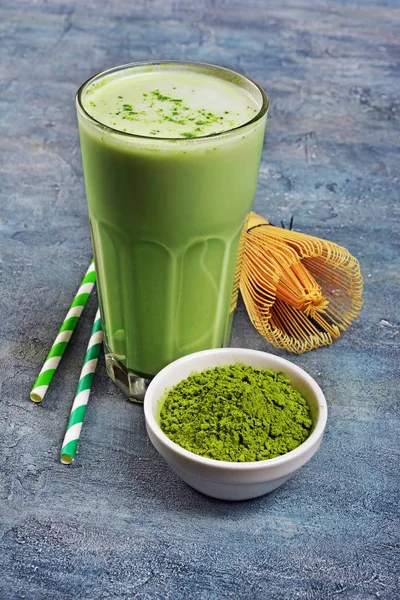 stock image Organic green matcha tea latte in glass and powder in white bowl with bamboo whisk and cocktail straw on blue concrete background