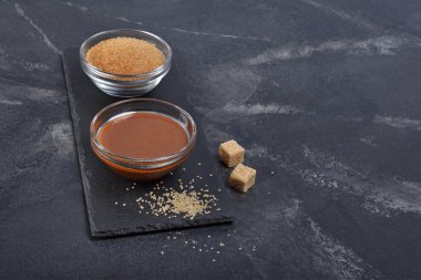 Ev yapımı tatlı karamel sos cam kase ve siyah arduvaz kurulu ve mermer arka plan kopya alanı ile kahverengi şeker kamışı