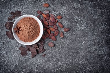 Kopya alanı koyu gri beton zemin üzerine şekerlemeler için madde olarak çikolata ve bütün kakao çekirdeği parçaları ile beyaz kase kakao tozu üstten görünüm