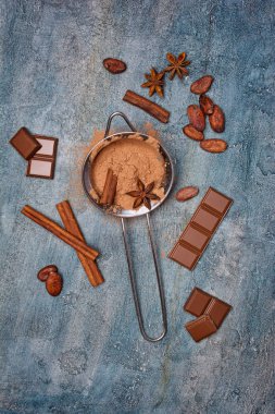 Çikolata, kakao çekirdekleri ve baharat olarak mavi beton zemin üzerine şekerlemeler için malzemeler ile süzgeç içinde kakao tozu üstten görünüm